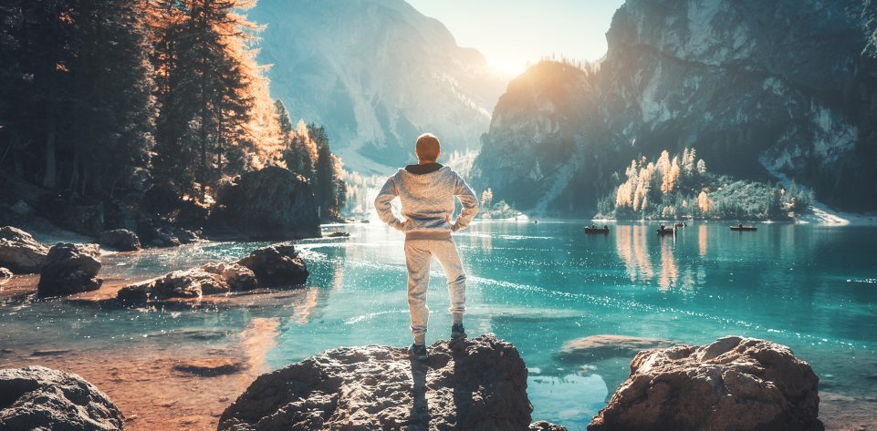 Standing young man on the stone on the coast of Braies lake at sunset. Autumn in Dolomites, Italy. Landscape with happy man, mountains, water with reflection, fall trees and blue sky in the evening